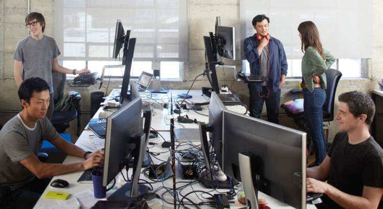 Open-plan office with several people working at computer stations while two colleagues stand and talk near standing desks.