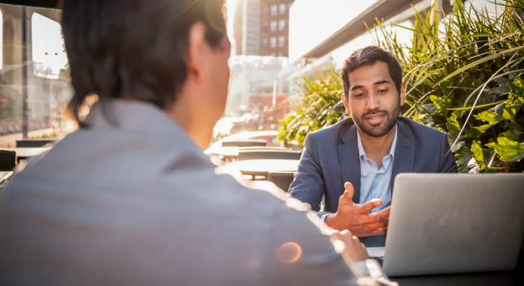 Two colleagues talking at an outdoor cafe table, with one person in a suit speaking and using a laptop in a sunny city setting.
