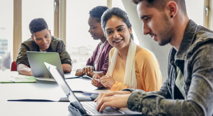 Group of students sitting together at a table with laptops, collaborating and smiling during a study session.