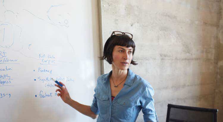 Person in a blue shirt with glasses resting on their head standing next to a whiteboard, pointing with a blue marker.