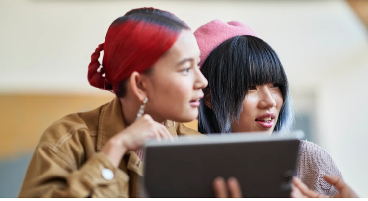 Two individuals seated closely indoors, looking at a tablet together; one wears a brown jacket and earrings, the other a pink beret and textured sweater, suggesting a collaborative or learning moment.