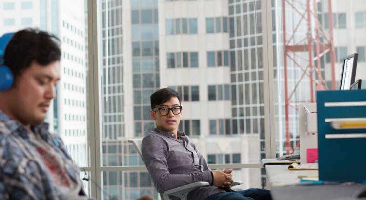 Two individuals in a modern office with large windows overlooking a cityscape; one wears blue headphones and a plaid shirt, the other wears glasses and a gray shirt, seated near desks with a monitor and file organizer.