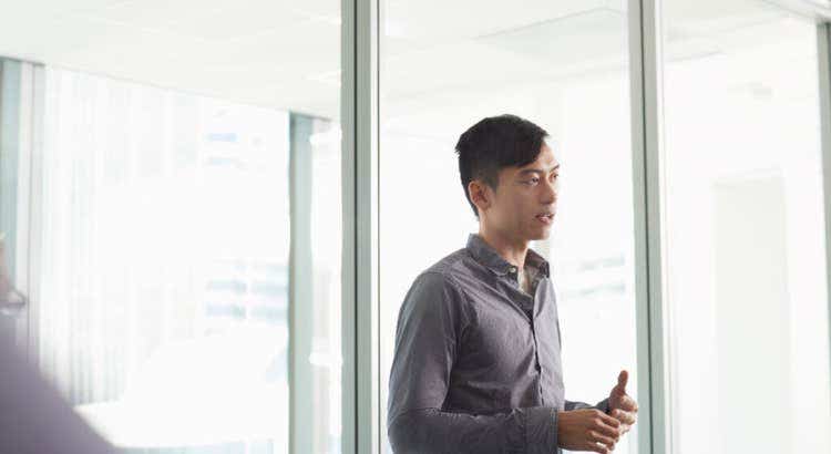 Person standing in a bright glass walled conference room, wearing a long sleeve shirt and gesturing with their hands as if giving a presentation or speaking to colleagues.