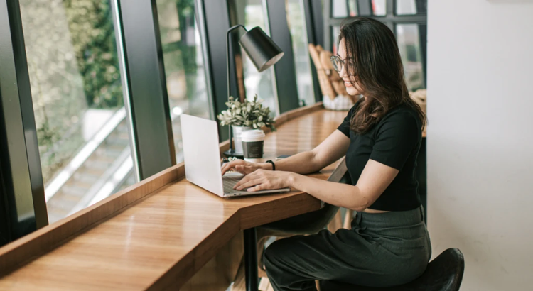 Person in casual attire working intently on a laptop at a wooden café counter beside a large sunlit window, with a desk lamp, takeaway coffee cup, and potted plant creating a cozy, modern remote work setup.