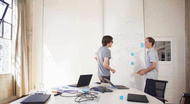 Two professionals engaged in a brainstorming session at a modern office whiteboard filled with diagrams and blue sticky notes, with a conference table in the foreground holding laptops, papers, and a phone, all bathed in natural light from large windows.