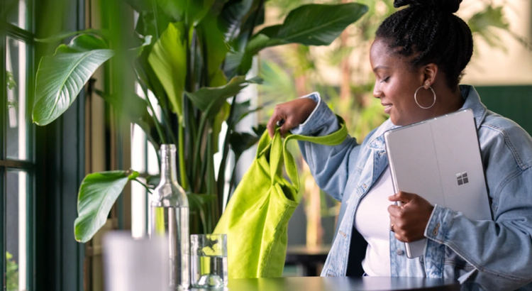 Person standing indoors near a table and lush green plants, holding a bright green cloth and a Microsoft Surface tablet. Dressed in a light blue denim jacket and white shirt, with hoop earrings and braided hair in a bun. The modern space features large windows, natural light, and minimalist decor with glassware on the table, suggesting a relaxed, creative environment.