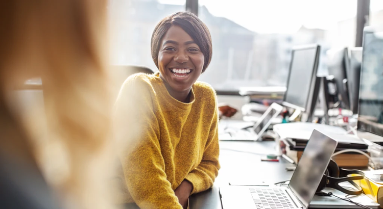 Person in a yellow sweater smiling during a conversation at a desk in a bright, open office. A laptop, papers, and office supplies are on the desk, with multiple monitors and desks visible in the background. Natural light from large windows creates a warm, collaborative atmosphere.