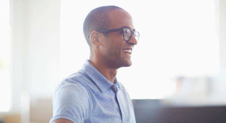 Person in glasses and light blue shirt seated indoors near a window, engaged in conversation in a bright office setting.