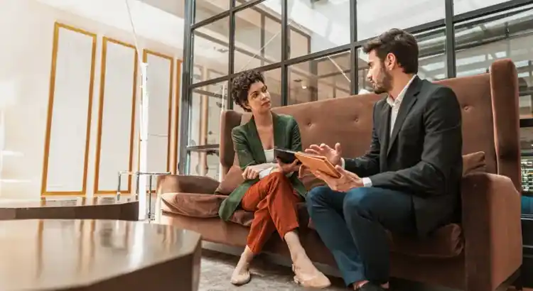 Photo of two individuals seated on a brown couch in a modern office with glass panels and gold-framed wall decor, engaged in a serious conversation with a notebook and tablet, suggesting a professional consultation or business meeting.