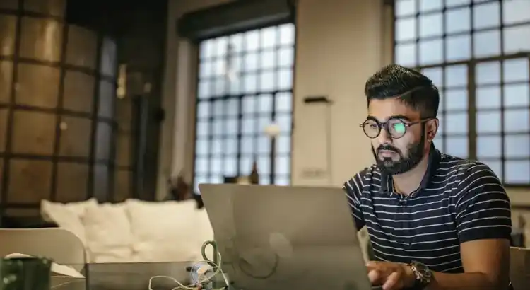 Photo of a person working on a laptop in a modern industrial-style room with large grid windows, exposed beams, and a white couch in the background, suggesting a focused home office or co-working space.