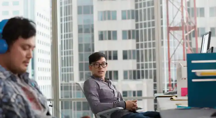 Photo of two individuals in a modern office with city views, one wearing blue headphones and focused on a device, the other seated near a desk with a monitor and papers, depicting a tech-enabled urban workspace.