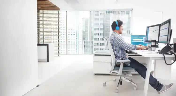 Person in blue headphones working at desk with dual monitors displaying charts in bright office with city view
