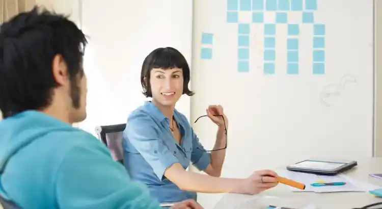 Two people discussing in front of whiteboard with blue sticky notes and dinosaur drawing in modern office