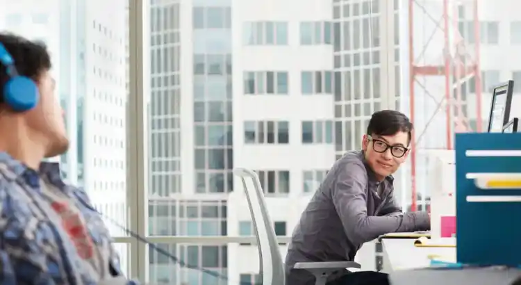 Two coworkers in office with city view, one in plaid shirt and headphones, the other smiling at desk with monitor