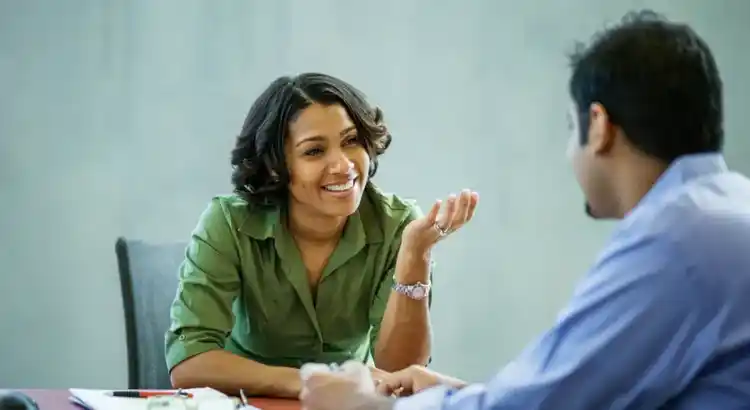 Two people seated at table in discussion, one gesturing expressively in green shirt, the other listening in blue