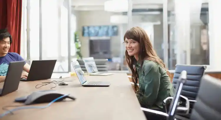 Two people using laptops at long conference table in bright office with glass-walled meeting room