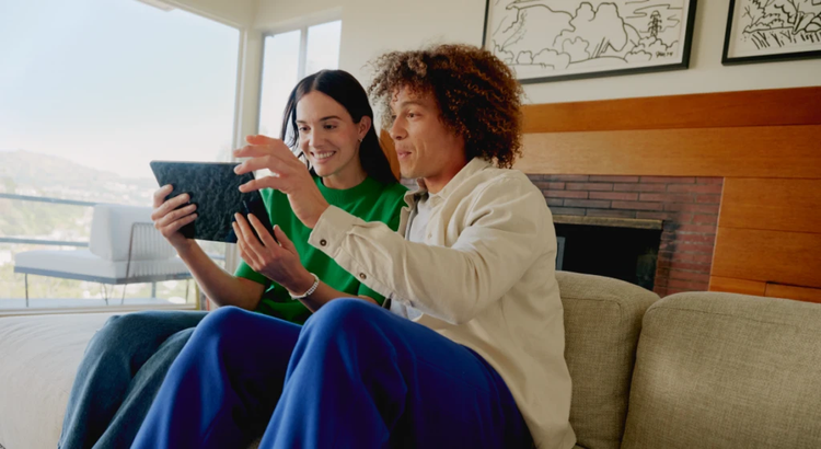 Two people sitting on a beige couch in a bright living room, interacting with a tablet near a fireplace.
