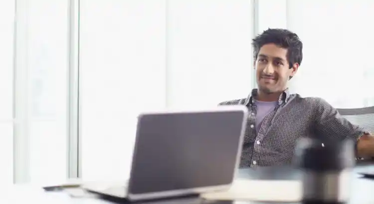 Person leaning back in a chair at a desk with an open laptop, notebook, and coffee cup in a sunlit office.