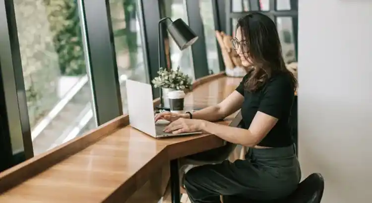 Casual remote work scene with laptop user in a sunlit cafe featuring coffee cup, plant, and desk lamp.