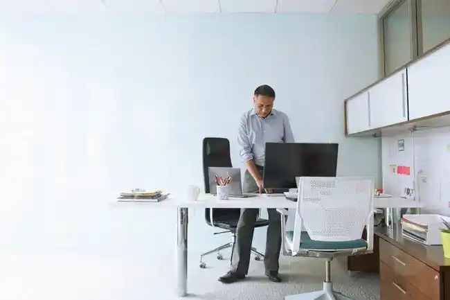 Modern office workspace with a person at a white desk, surrounded by chairs, supplies, and a bulletin board.