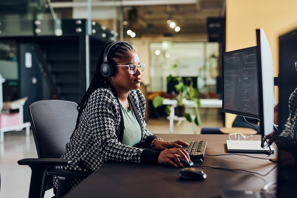 lack female software engineer wearing headphones and glasses, working on code at a computer in a modern office. Focus on women in tech and inclusive work environments.