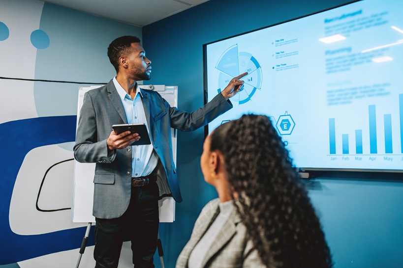 A business professional presents data visualizations on a large screen to a colleague, demonstrating Microsoft's commitment to clear communication and data accessibility for all stakeholders.