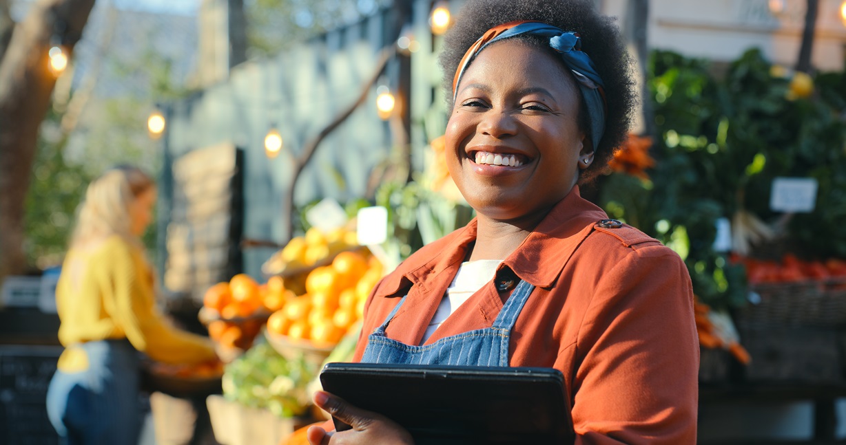 A smiling Black woman wearing an apron and holding a tablet stands in front of a market stall filled with fresh produce. Focus on small business ownership, local markets, and technology adoption.