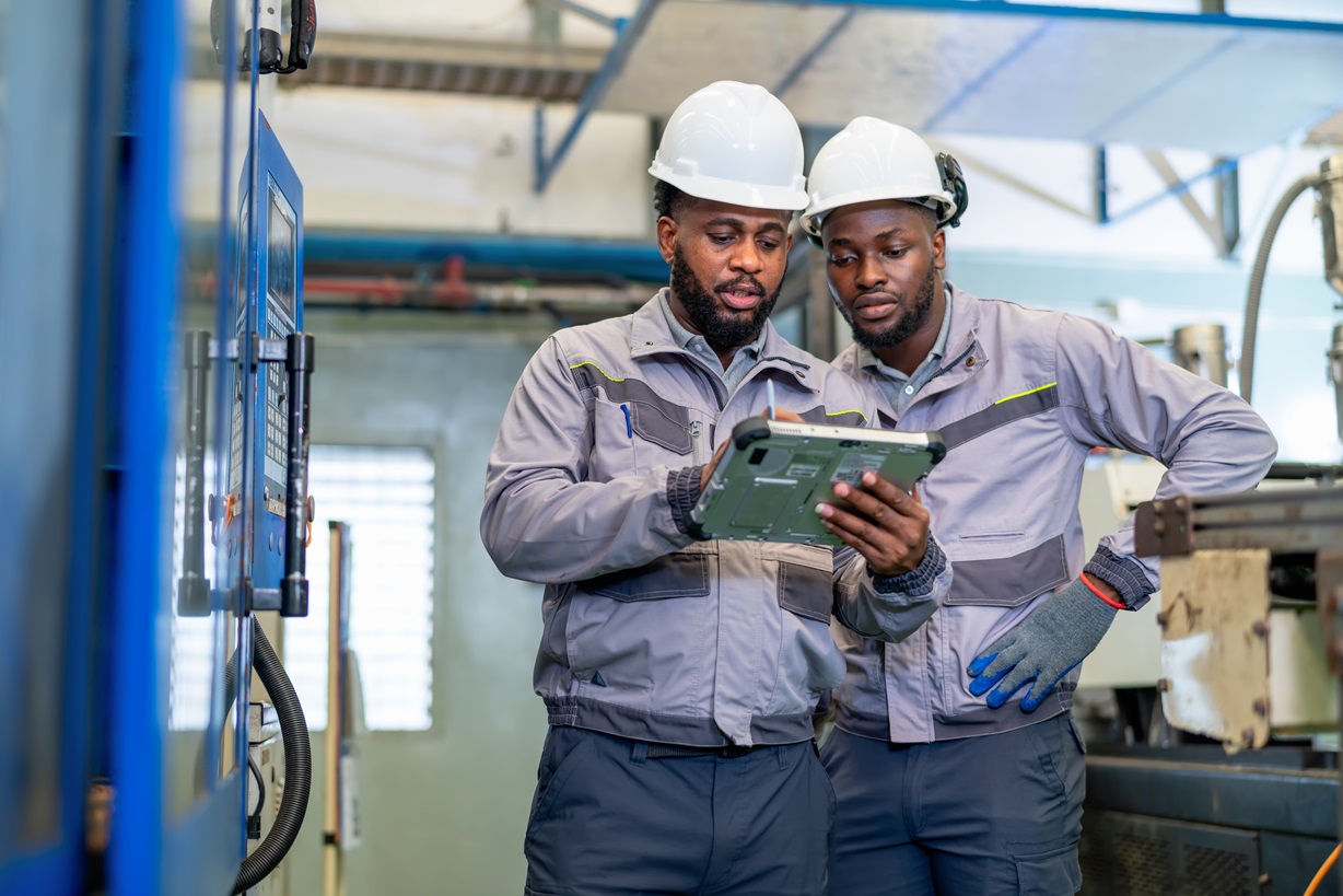 Two Black male factory workers wearing hard hats and safety gear examine a tablet while standing near industrial machinery. Focus on teamwork, technology in manufacturing, and worker safety.