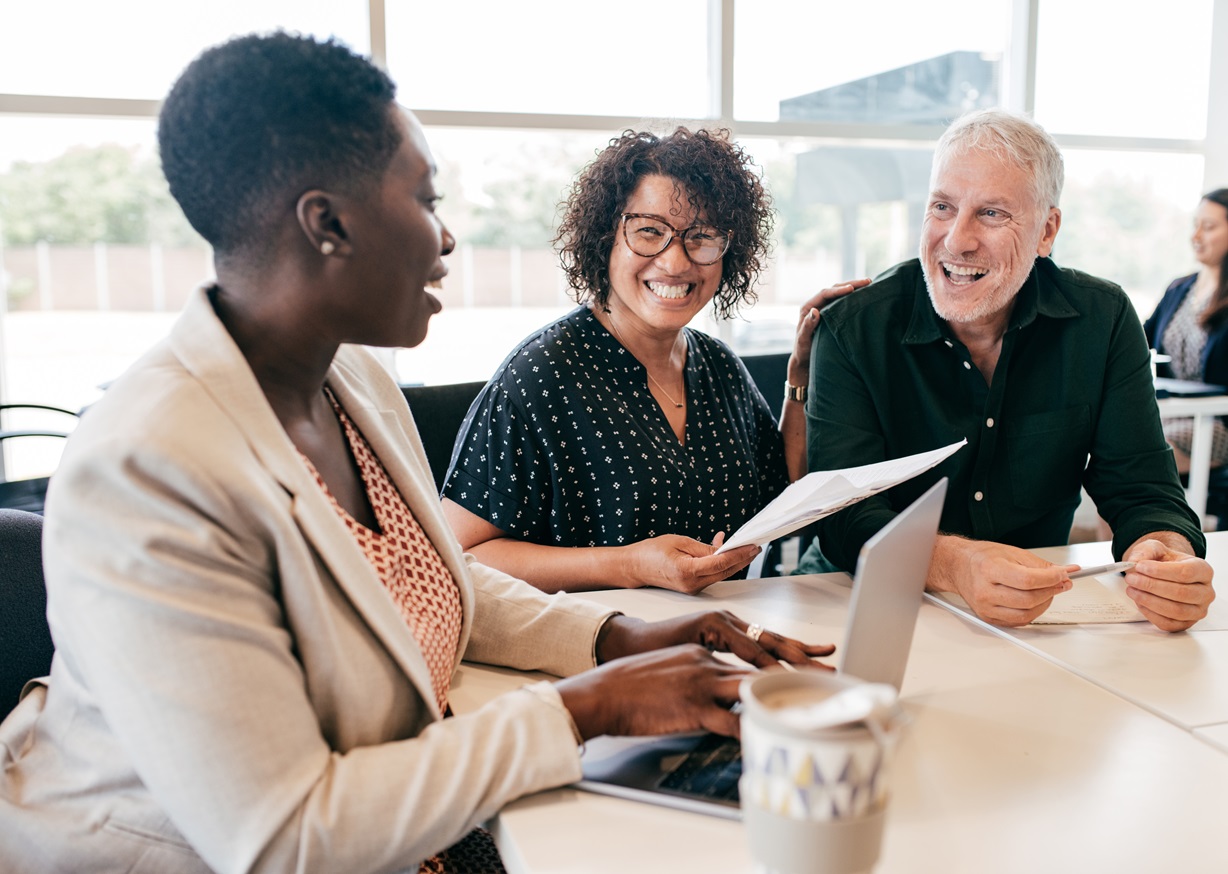 A diverse group of business professionals, including a Black woman, a white woman, and a white man, meet at a table with a laptop and documents. Focus on collaboration, diversity in the workplace, and business meetings.