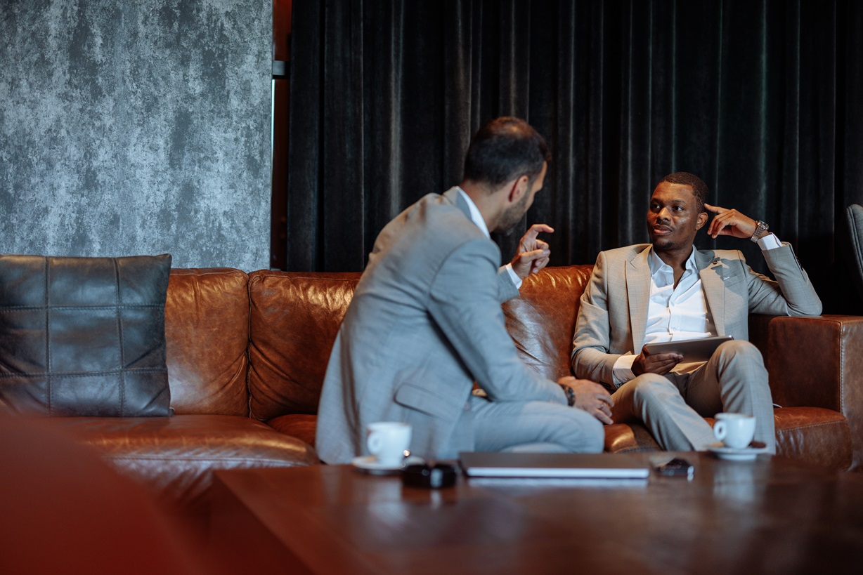 Two Black men in suits engaged in a business discussion on a leather couch. It represents leadership, collaboration, and professional development, emphasizing diversity in corporate settings.