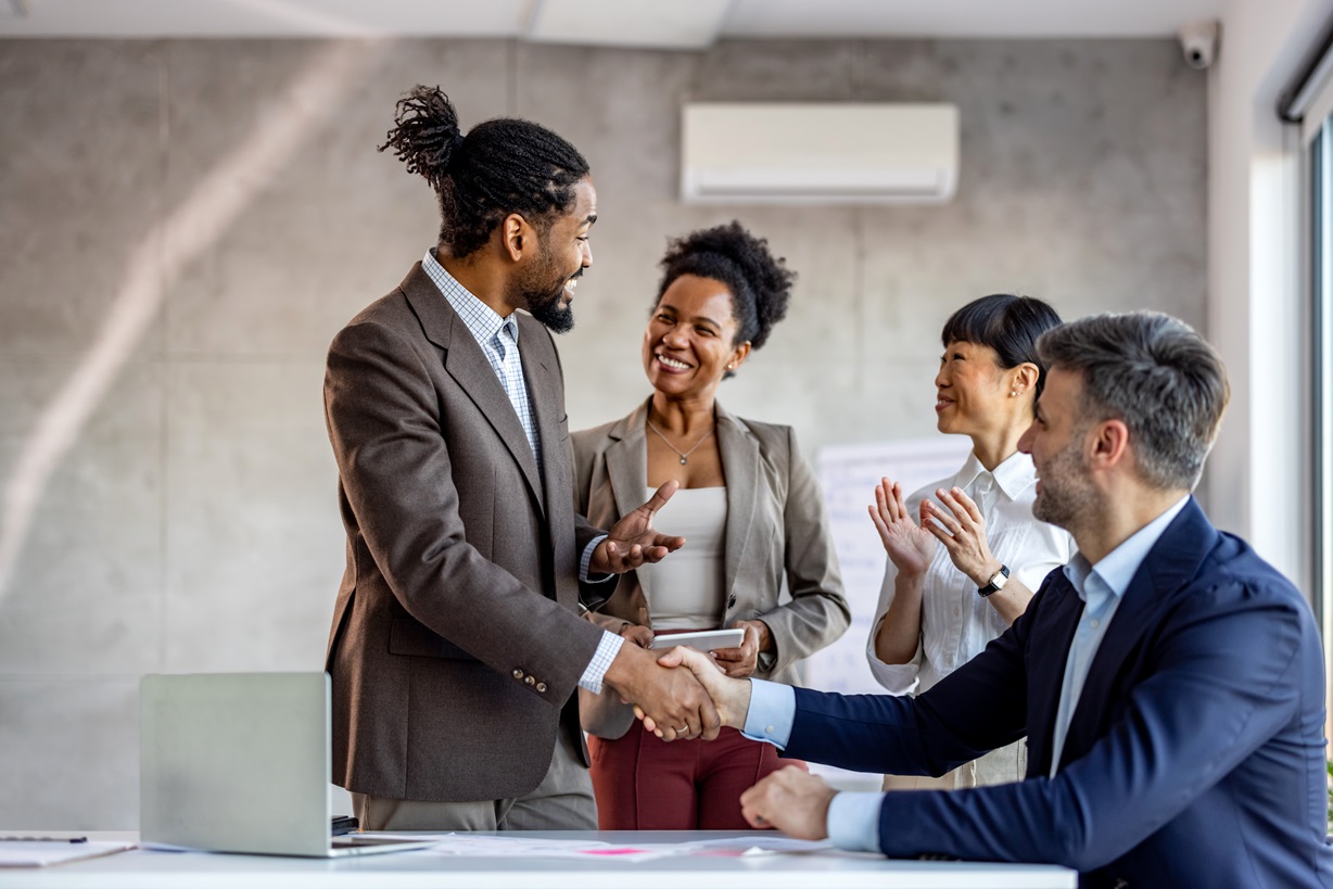 A diverse group of professionals celebrating a successful deal with a handshake and applause. It represents teamwork, achievement, and the value of diversity and inclusion in a collaborative work environment.