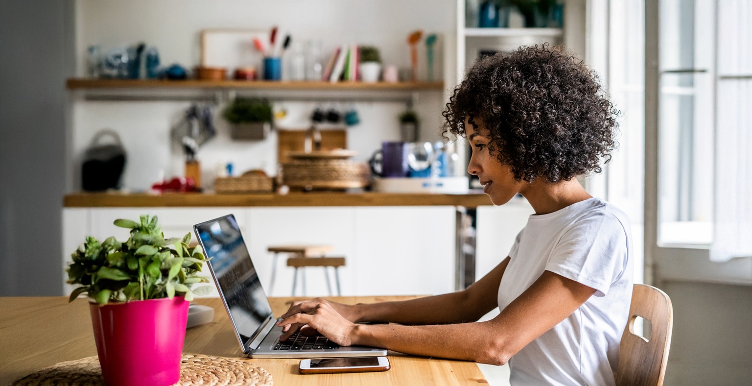A young Black woman with curly hair works on a laptop at a kitchen table. A plant in a pink pot sits beside her. The scene suggests flexible work arrangements and a focus on employee well-being in a comfortable environment.