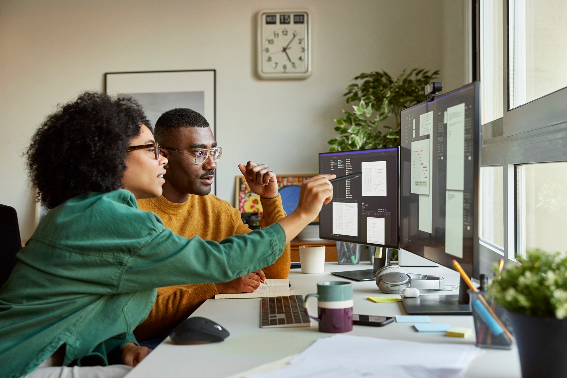 A Black man and woman work together on a coding project, using multiple computer screens. The woman is pointing at the code. It represents teamwork, technical skills, and the importance of diversity in STEM fields.
