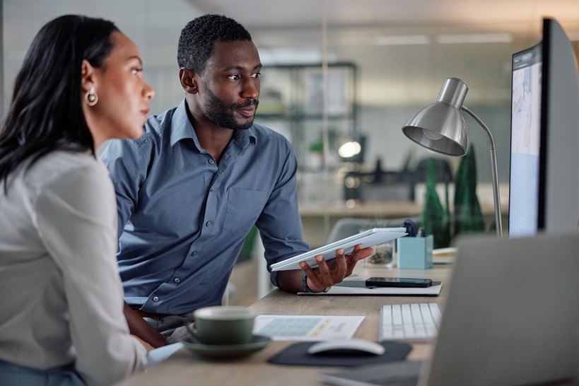 Two Black professionals, a man and a woman, collaborate in an office setting. The man is holding a tablet and gesturing towards a computer screen, indicating a discussion about data or design