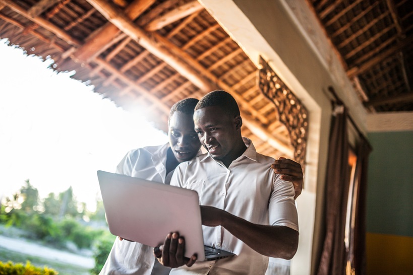 Two young Black men are looking at a laptop together on a porch or balcony. One has his arm around the other's shoulder. It suggests collaboration, possibly in a relaxed or informal setting, and highlights the importance of mentorship or peer support.