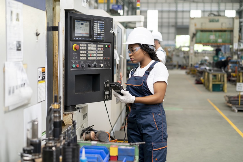 A Black female engineer wearing safety gear, operating a CNC machine in a manufacturing facility. This highlights the role of women in STEM and the use of technology for efficient and sustainable production.
