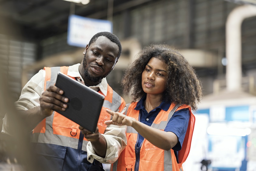 Two Black manufacturing workers in safety vests, collaborating with a tablet in a factory setting. This showcases teamwork and the use of digital tools for optimizing processes and promoting sustainability in manufacturing.