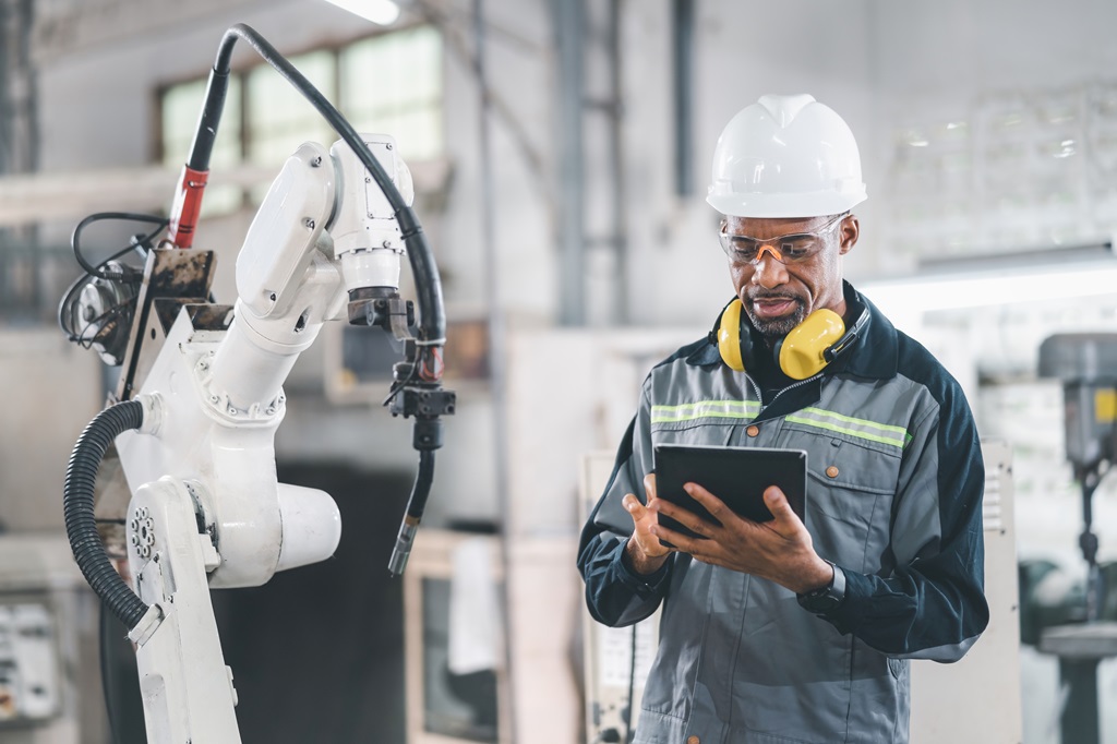 A Black male engineer wearing safety glasses, a hard hat, and ear protection, uses a tablet while standing next to a robotic arm in a factory. It highlights the intersection of technology and skilled labor, showcasing innovation in manufacturing.