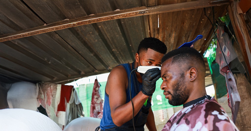 A Black barber, wearing gloves, gives a haircut to a client. It represents entrepreneurship, small business ownership, and the provision of personal services.