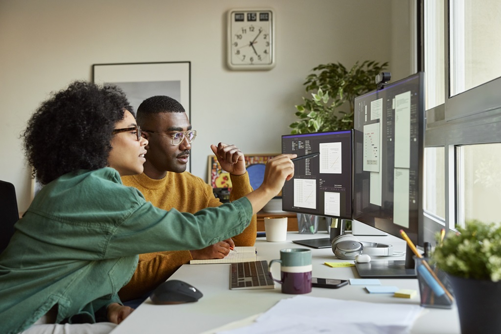 A Black man and woman work together on a coding project, using multiple computer screens. The woman is pointing at the code. It represents teamwork, technical skills, and the importance of diversity in STEM fields.