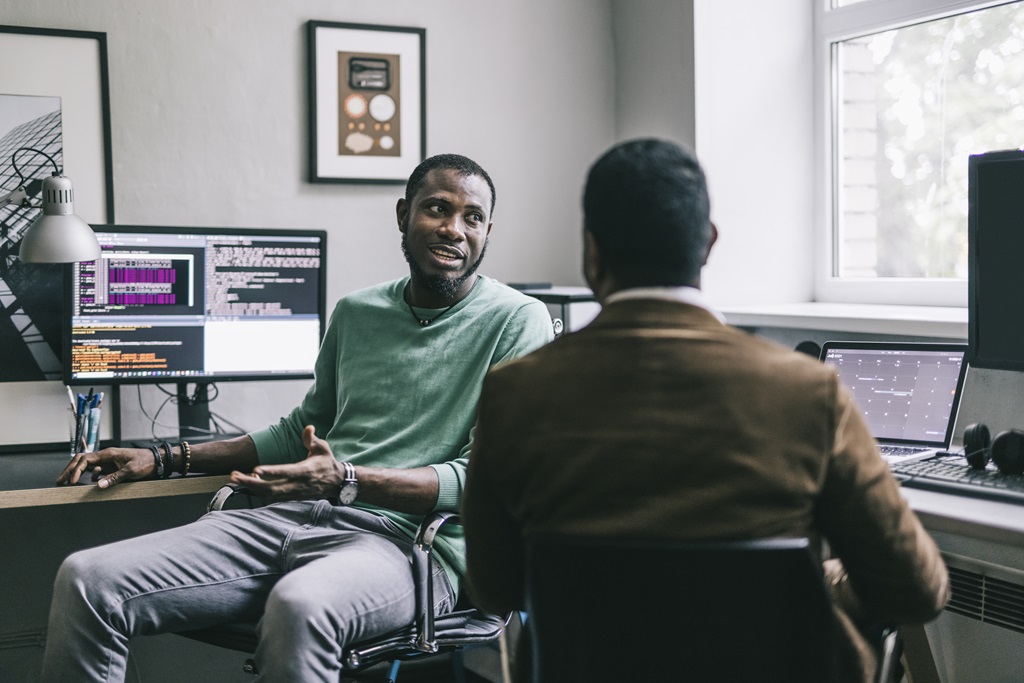 Two business professionals, one Black man in a green shirt and gray pants, and another Black man in a brown suit, discussing business in an office setting with multiple computer screens displaying code. Focus on collaboration and diverse representation in tech.