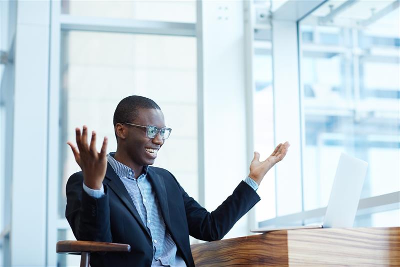 A young, enthusiastic Black professional celebrating a success while working on a laptop in a bright, modern office. It represents innovation and achievement in a tech-driven, environmentally conscious workplace.