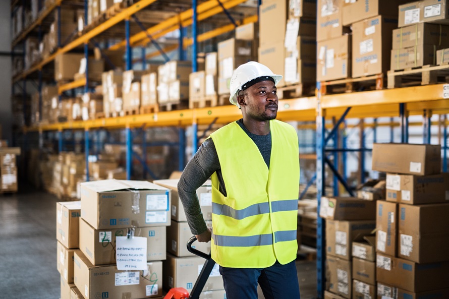 A warehouse worker wearing a hard hat and safety vest stands in a large warehouse filled with boxes. Focus on safety and efficiency in warehouse operations.