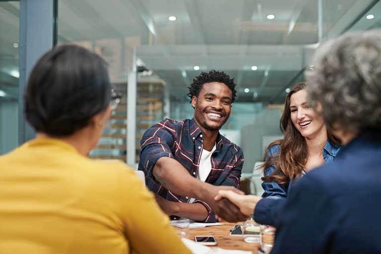 A Black man in a meeting shakes hands with a woman across a conference table. Other colleagues are present, suggesting a successful negotiation or partnership. It represents positive workplace relationships and successful business outcomes