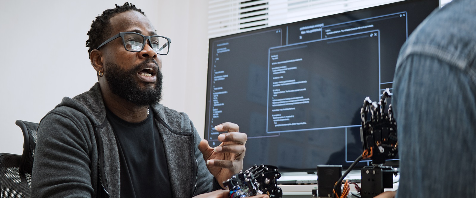 A Black man with glasses and a beard explains a robotic hand to a colleague. Code is displayed on a large monitor in the background. The scene highlights innovation and collaboration in technology, emphasizing diversity and inclusion in STEM fields.