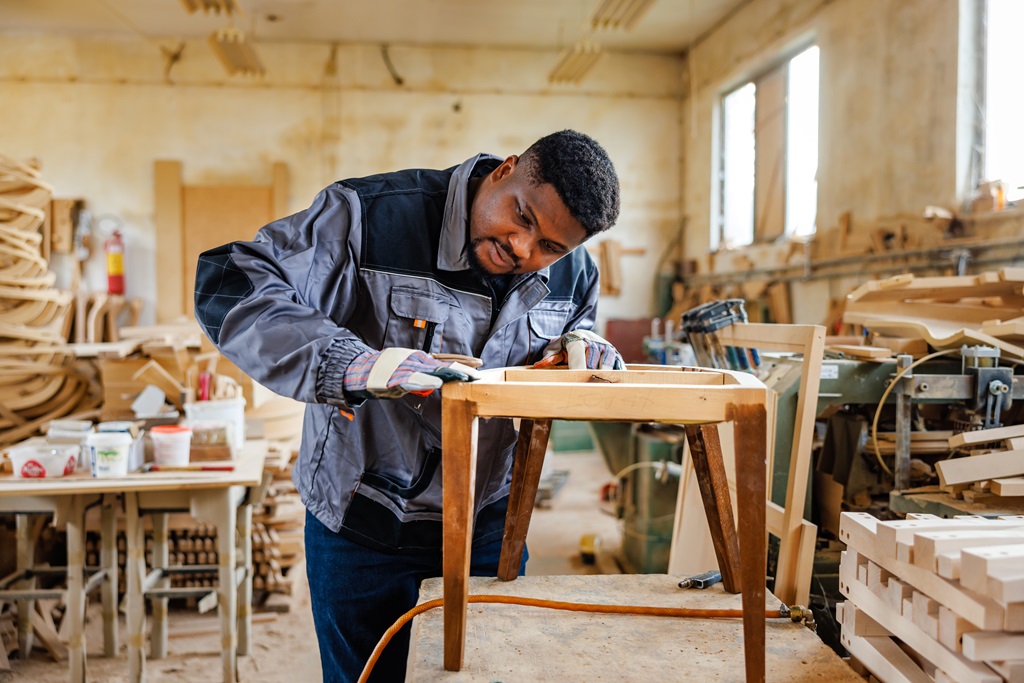 A Black man in a workshop sands a wooden chair. Woodworking tools and materials are visible in the background.