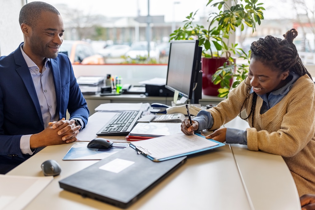 A Black woman signs a document at a desk while a Black man in a suit watches with a smile. A computer and office supplies are on the desk.