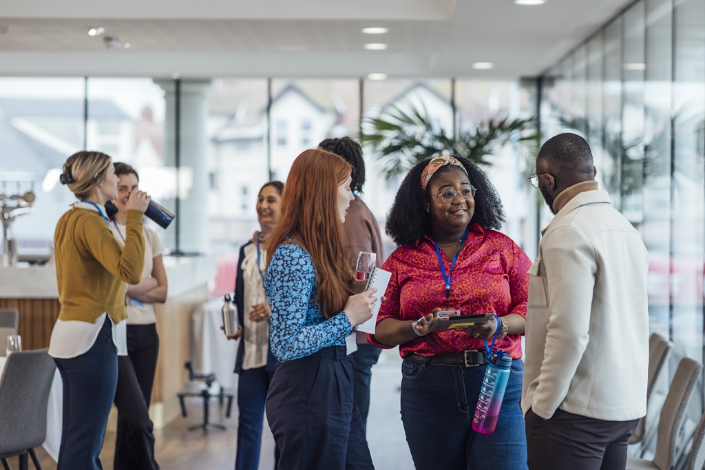 A diverse group of professionals networking at a conference. A Black woman in a red shirt holds a tablet and speaks with a Black man. Other attendees are visible in the background.