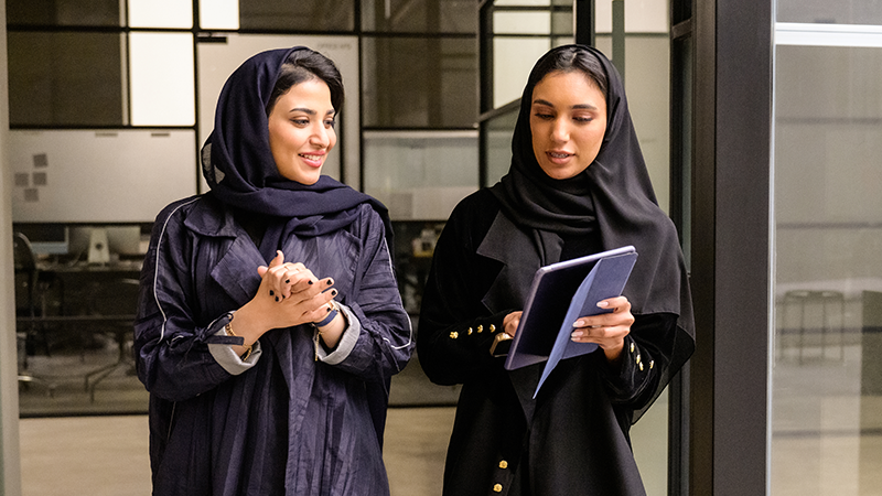 Two women walking while one woman has a smile on her face and the other woman showing her something on the tablet