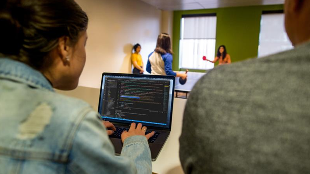 A woman sitting infront of a laptop side by side with a man, while three people play pingpong facing them